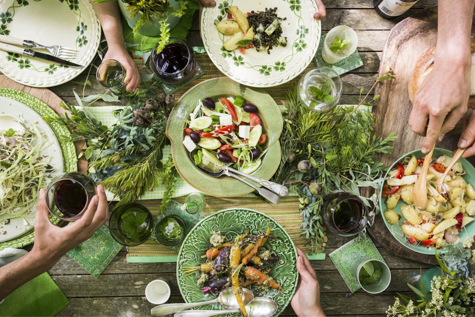 An outdoor meal with colorful salads, pasta, and roasted vegetables on decorative green plates, surrounded by fresh herbs. Hands are reaching in, sharing the food.